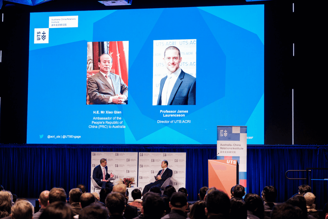 Two men in discussion at Australia-China Relations Institute event beneath plasma with two photos